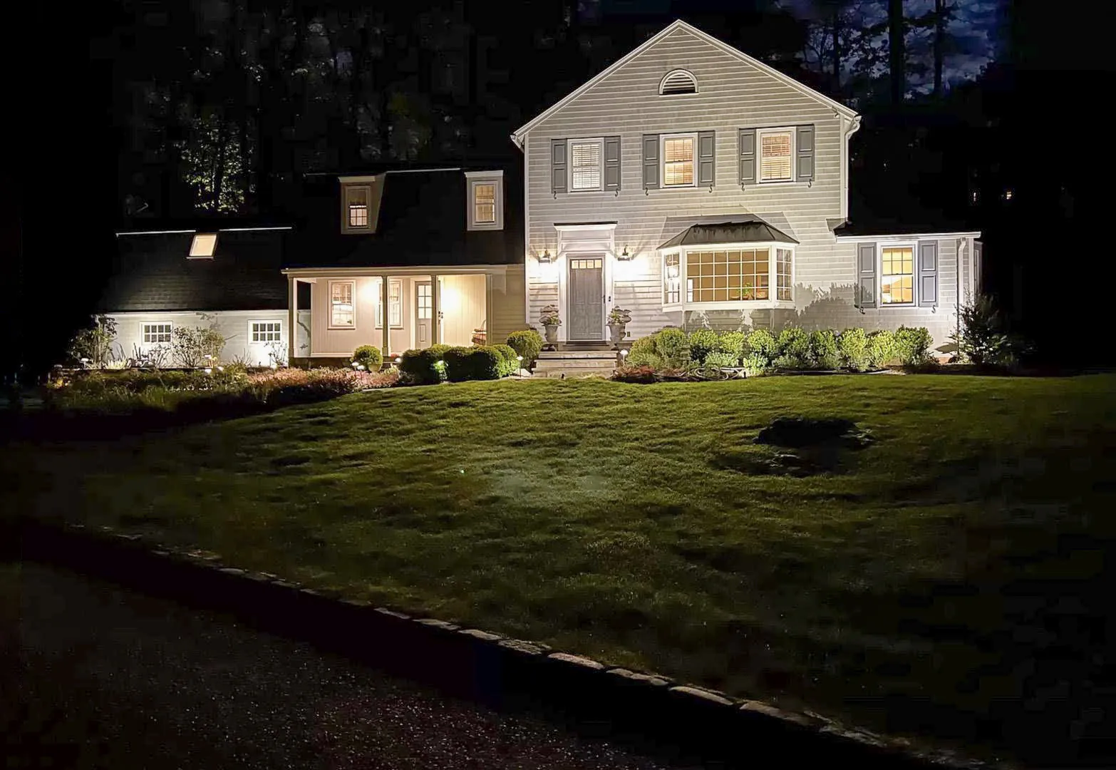 Shingled colonial lit at night by landscape lighting