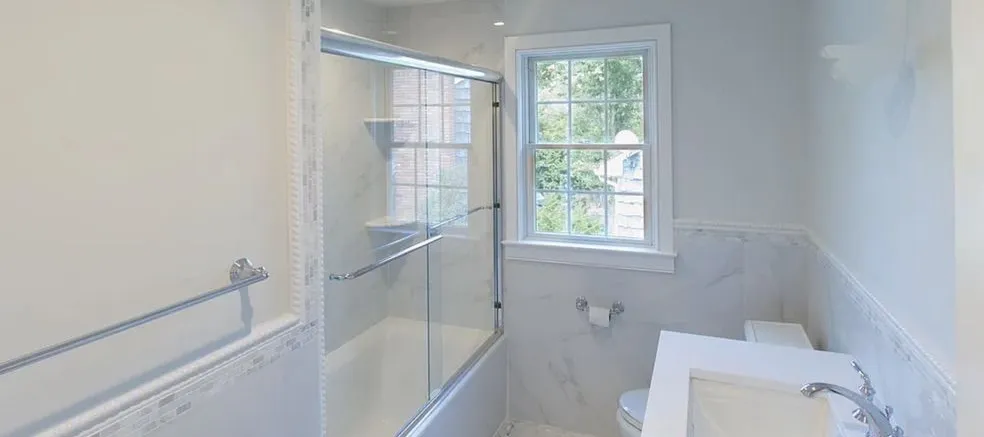 Marble bathroom with a glass tub enclosure, tiled wainscot and a window above the vanity
